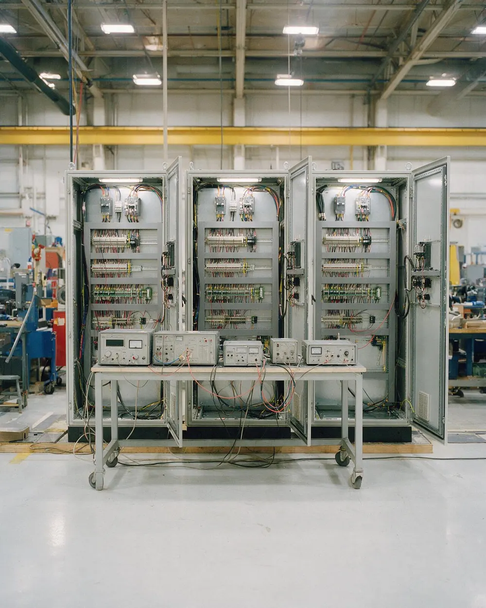 Three open control cabinets side by side in a workshop test bay, with a bench of test equipment in front